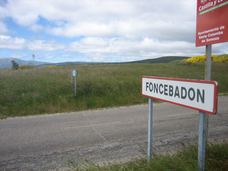 Ortsschild „Foncebadón“ an einer Landstraße, dahinter Wiesen und Hügel